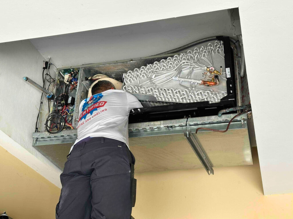 A Certified Heating and Cooling technician stands on a ladder and repairs the exposed internal parts of a ceiling-mounted AC unit.