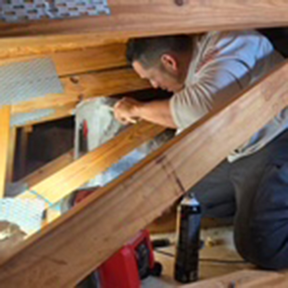A person installs insulation between wooden beams in an attic, with tools and materials scattered nearby to boost AC efficiency.