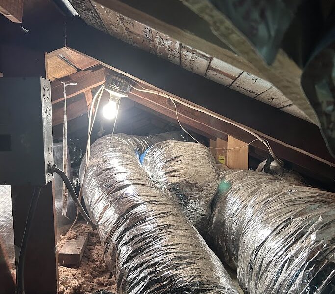 Las vigas expuestas, el aislamiento y los conductos flexibles en un ático del suroeste de Florida revelan el cableado y una bombilla encendida, lo que pone de relieve la necesidad de sellar los conductos.