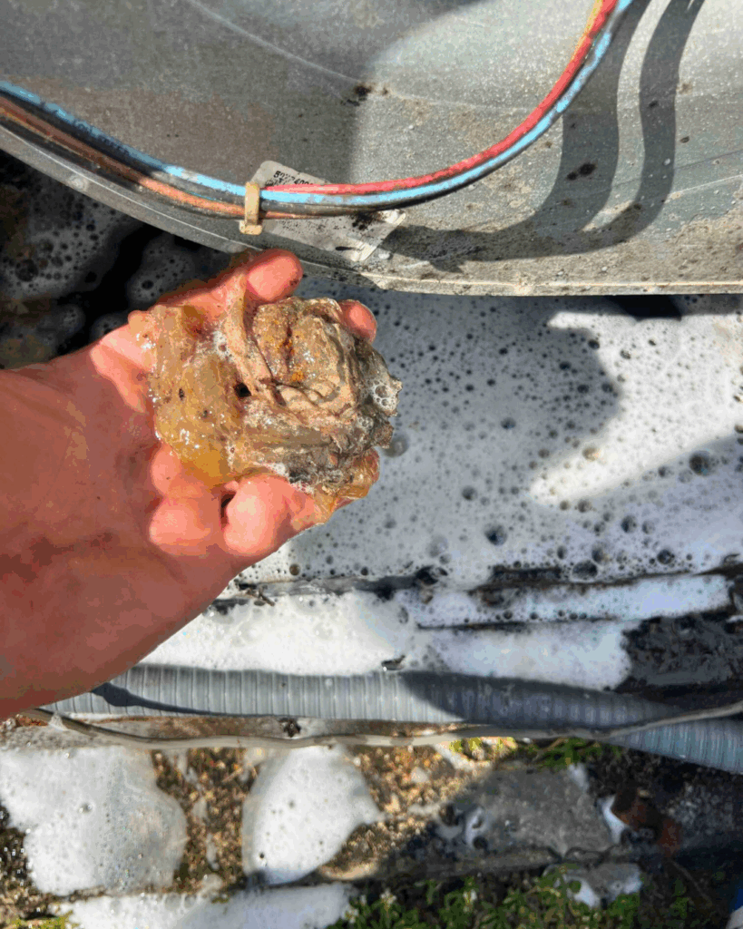 Una mano agarra un trozo sucio y gelatinoso de un equipo de climatización; en el fondo se ven espuma de jabón y cables.