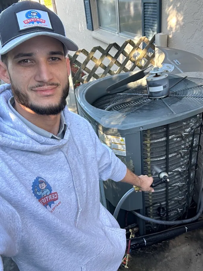 A man in a gray hoodie and cap removes the cover from an outdoor air conditioning unit and uses a tool to perform HVAC maintenance.