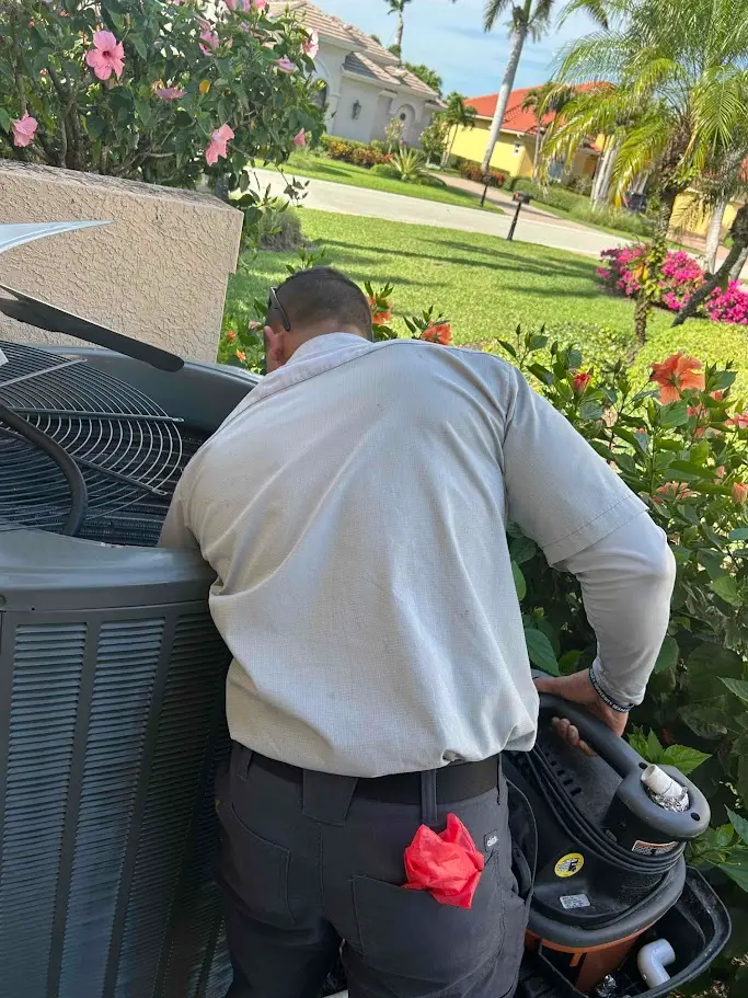 A technician in a gray shirt tunes up an outdoor air conditioning unit next to a garden with blooming flowers.