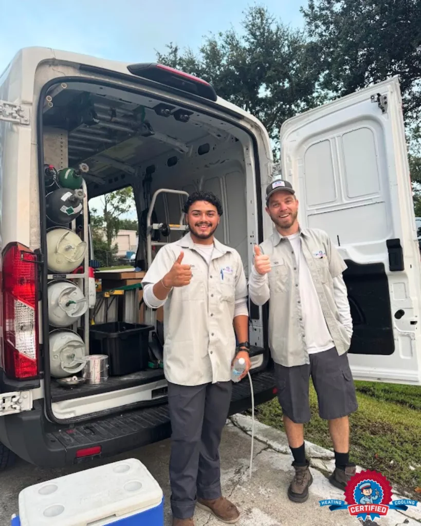 Two uniformed workers stand by an open service van, smile, and give thumbs up. Residential AC equipment fills the van. A technician badge appears in the corner.