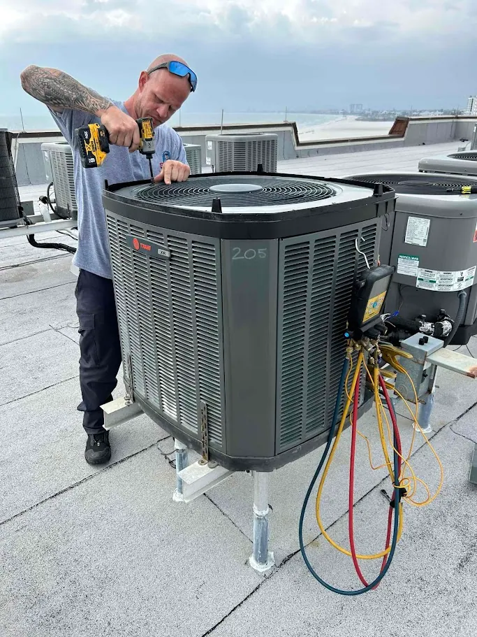 A technician uses a power drill to service a rooftop air conditioning unit equipped with diagnostic gauges.