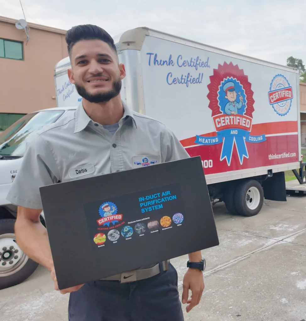 A uniformed technician holds an in-duct air purifier box beside a Certified Heating and Cooling truck to improve your indoor air quality.