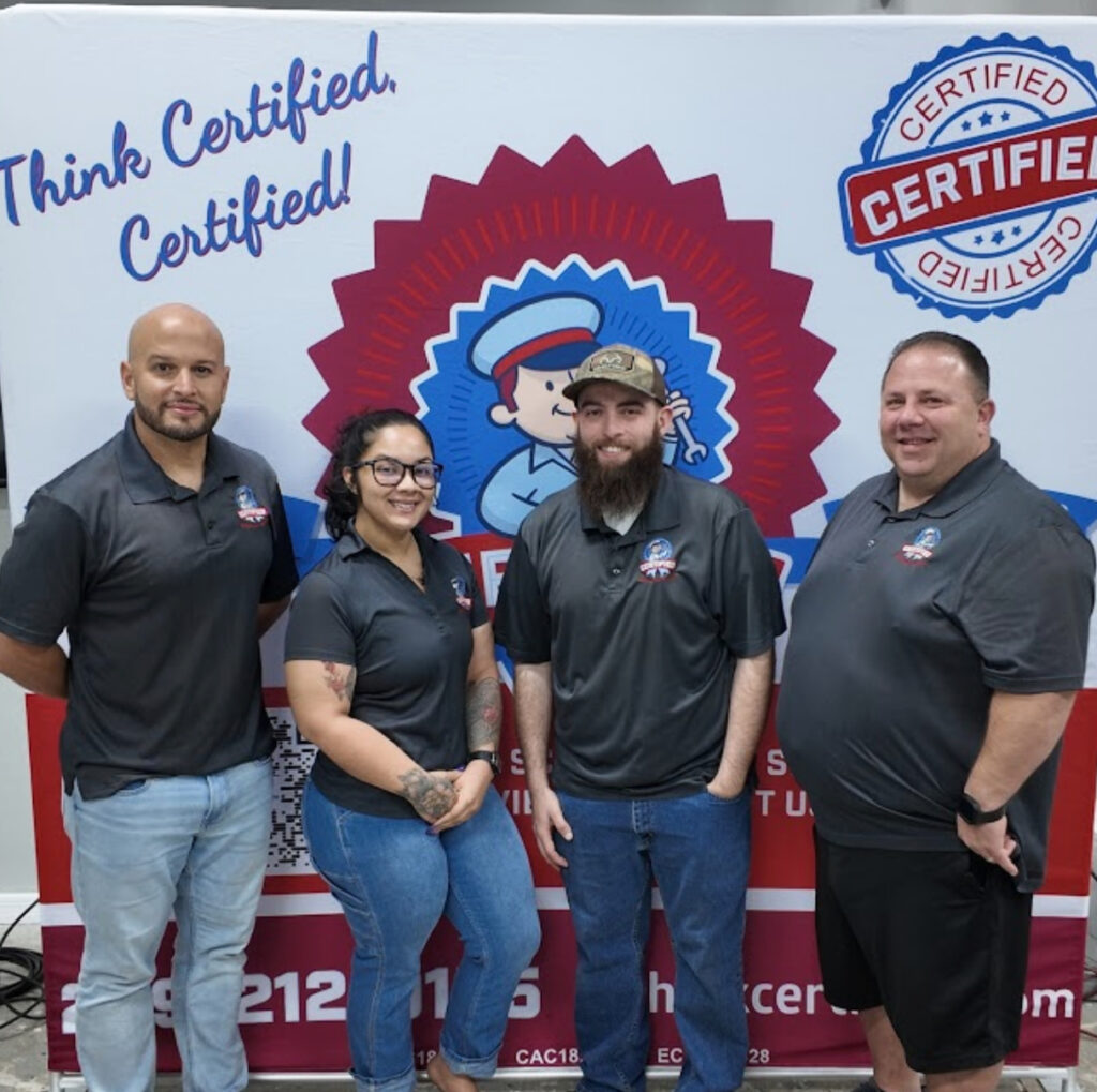 Four people in matching company shirts pose before a display reading "Think Certified. Certified!" with a cartoon logo and badges.