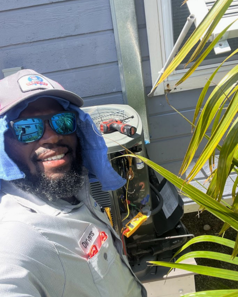 A technician in sunglasses, a hat, and blue cloth repairs an outdoor HVAC system beside a house on a sunny day, tools on the unit.