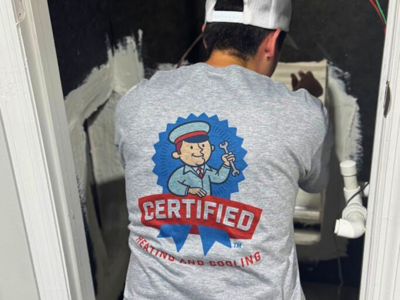 A person in a "Certified Heating and Cooling" shirt repairs equipment in a cramped, unfinished room with exposed wires and pipes.
