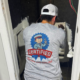 A person in a "Certified Heating and Cooling" shirt repairs equipment in a cramped, unfinished room with exposed wires and pipes.