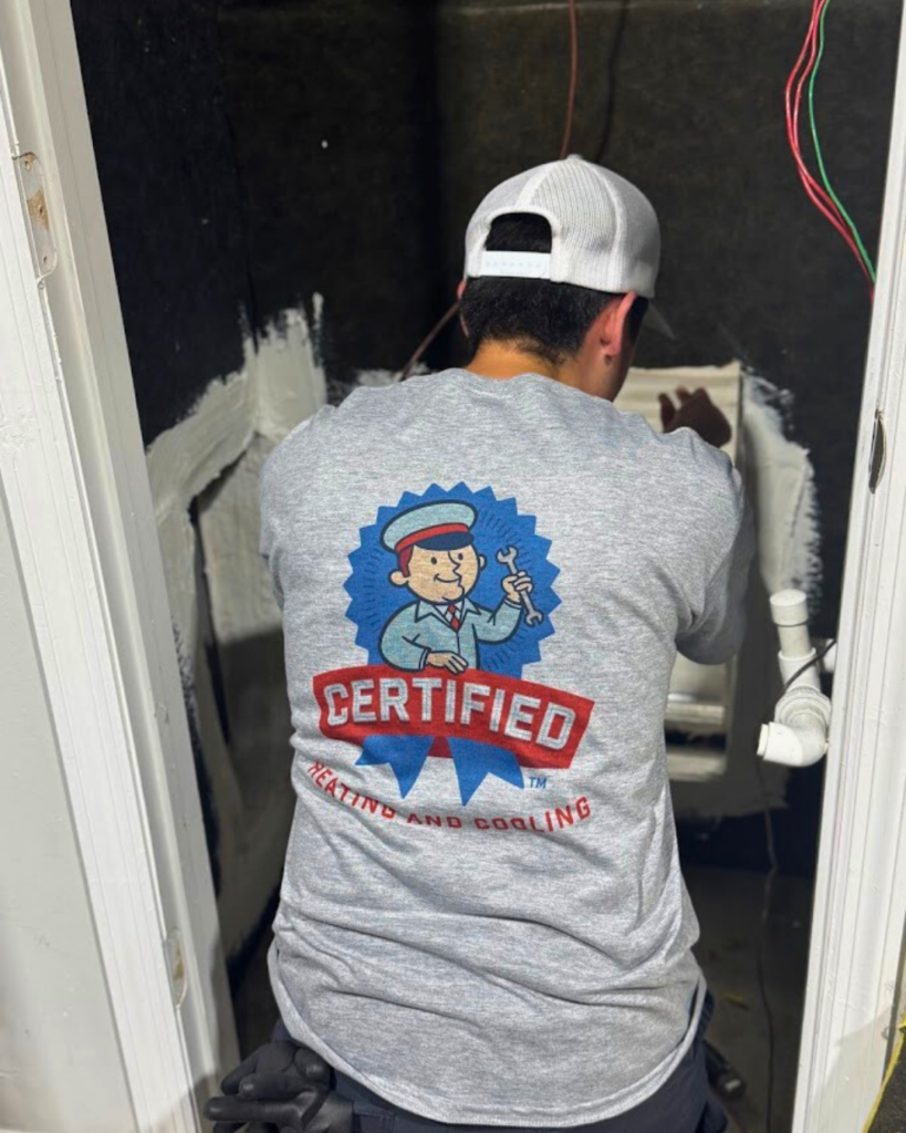 A person in a "Certified Heating and Cooling" shirt repairs equipment in a cramped, unfinished room with exposed wires and pipes.